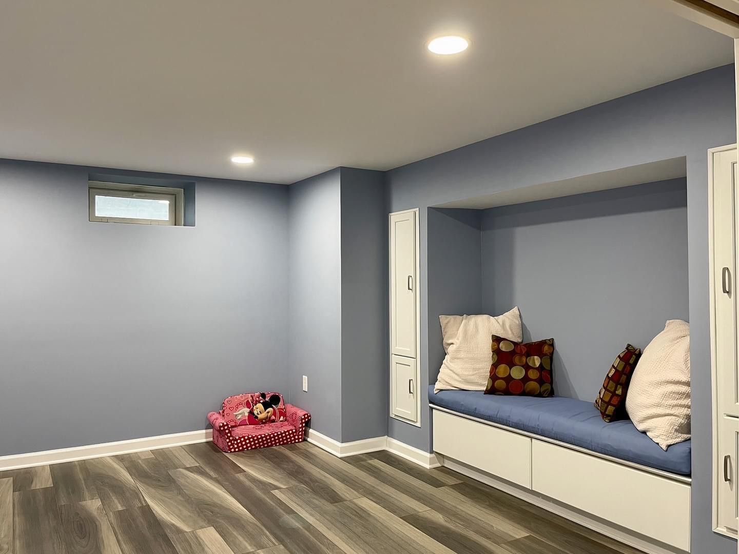 A cozy room with light blue walls, gray wood-look flooring, a window, a small pink chair, and a white bench seat with pillows.