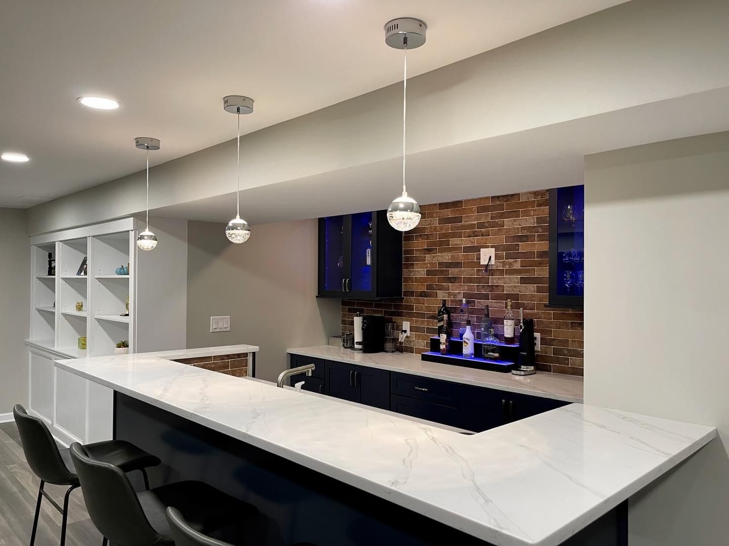A modern home bar with white marble countertops, navy cabinetry, pendant lighting, and a rustic brick-style backsplash.
