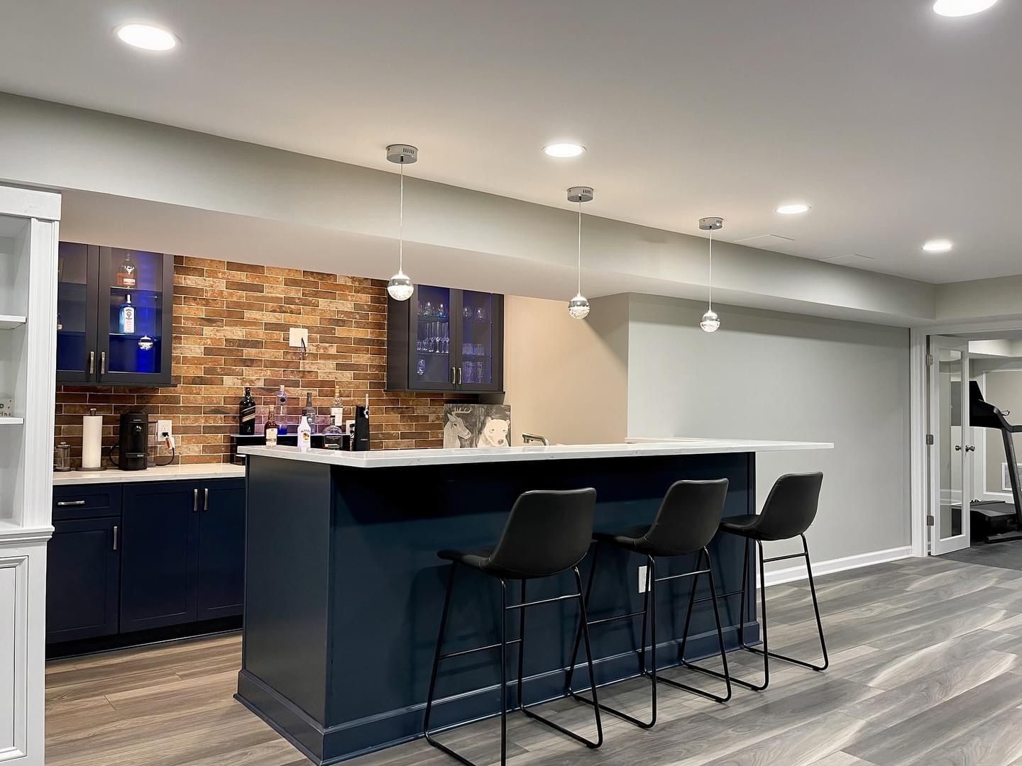 A basement bar with a dark blue island and stools, a stone backsplash, blue-lit cabinets, and wood-look flooring.