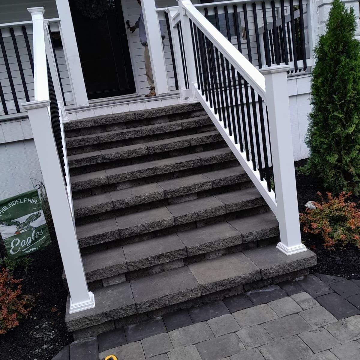 Stone steps lead up to a porch with white railings and black balusters, bordered by mulch and greenery.