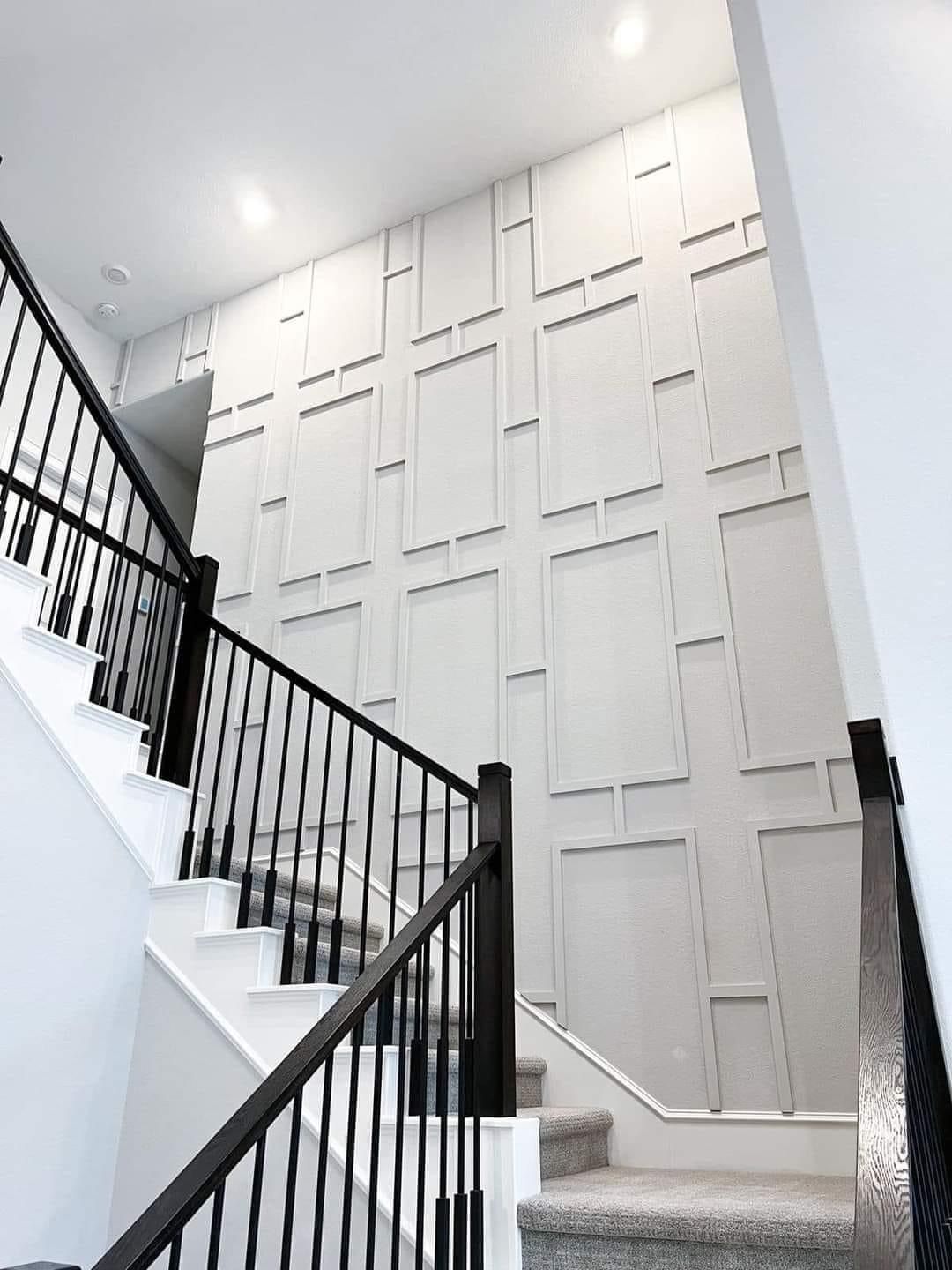 A staircase with a black railing, carpeted steps, and a floor-to-ceiling white accent wall with rectangular molding.