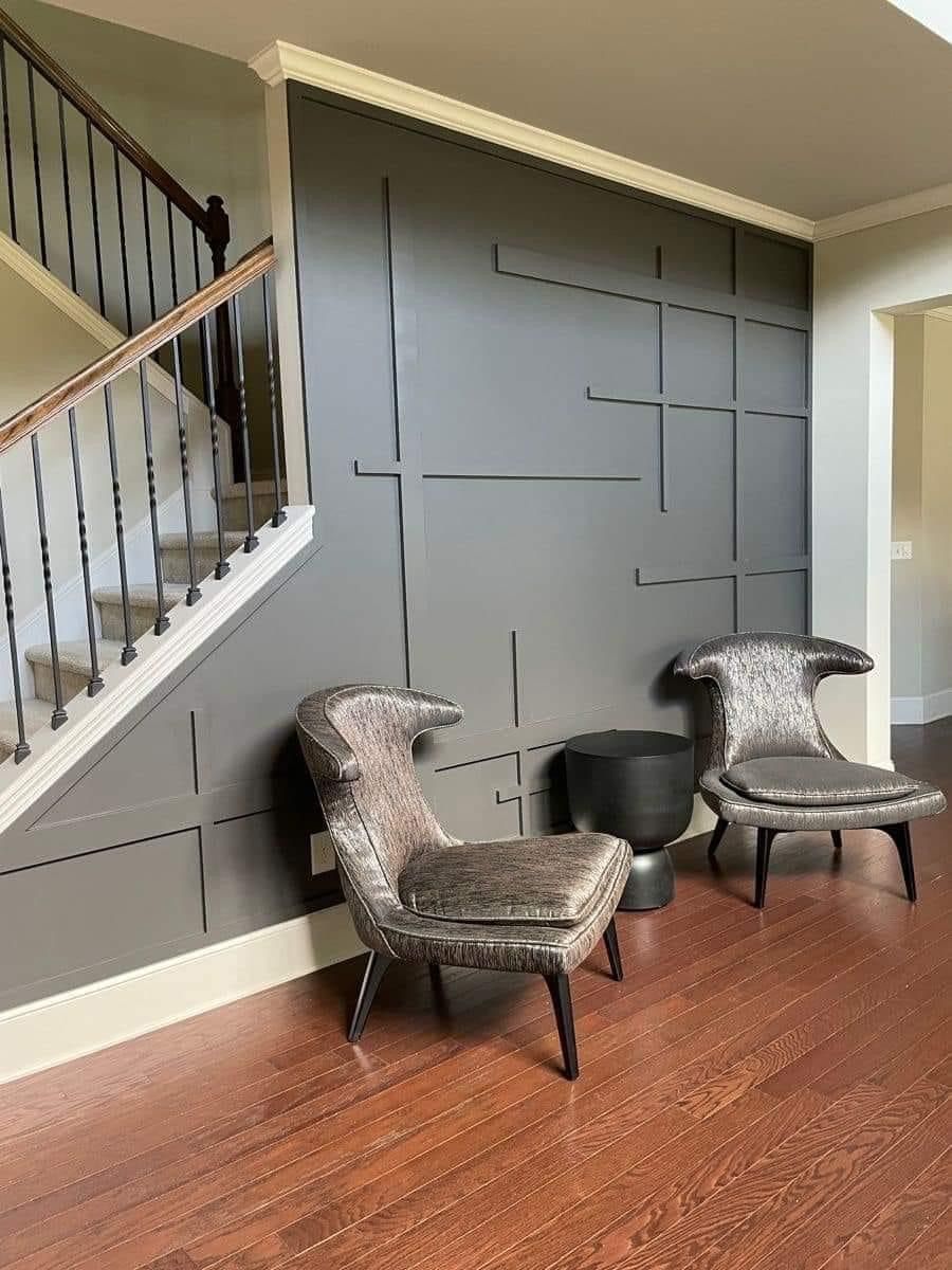 Two gray velvet accent chairs sit on a wood floor next to a staircase wall featuring a geometric, dark gray panel design.