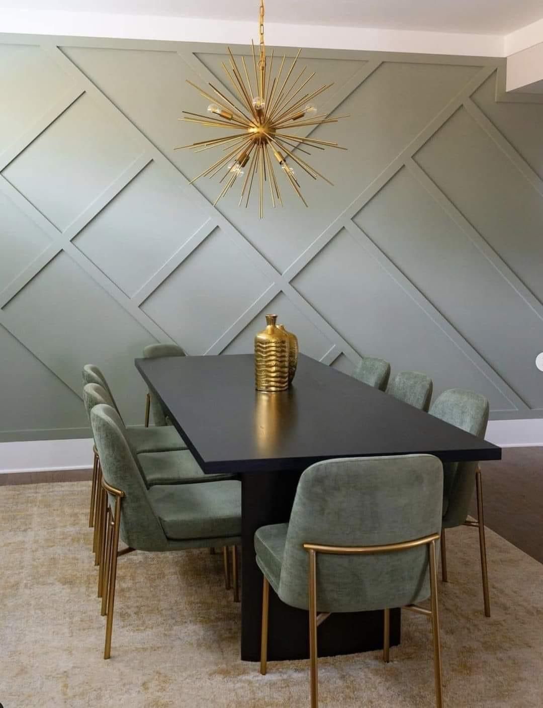 A dining table with sage chairs sits before a sage green wall with a large, geometric, raised-panel wood design.