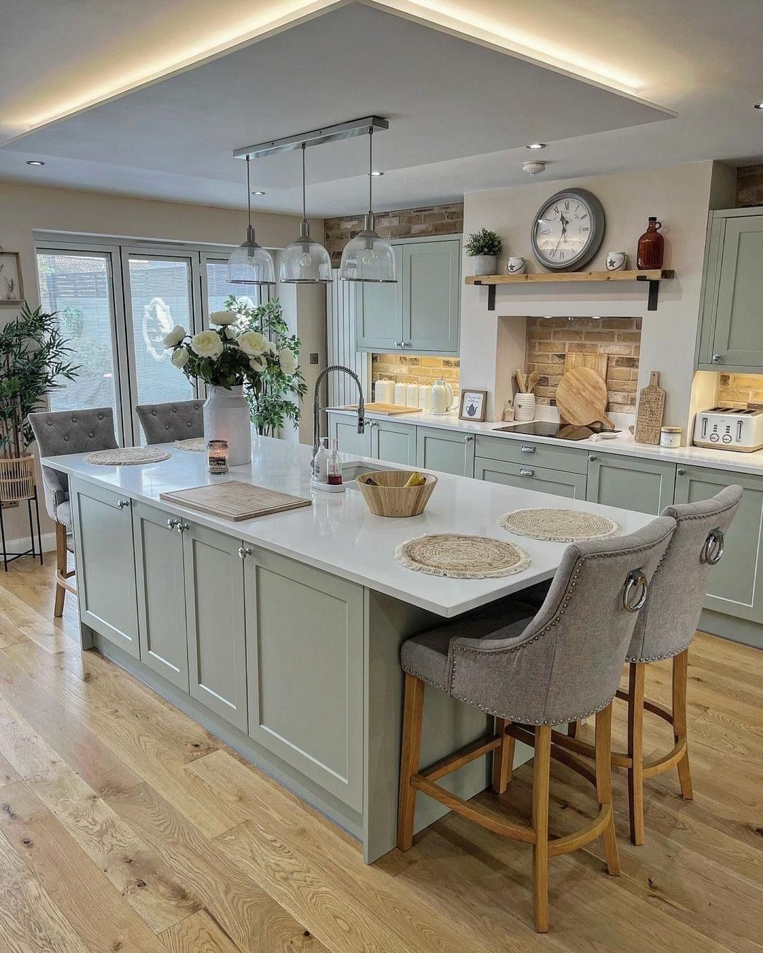 A modern, open-plan kitchen with a large sage green island, white countertops, bar stools, and warm wood flooring.