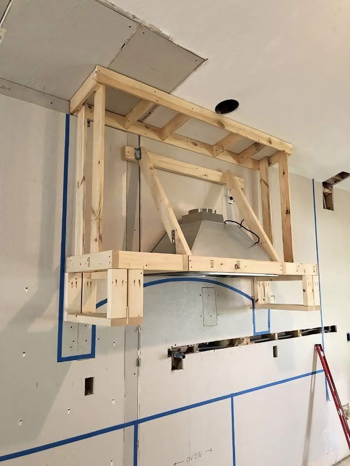 A wooden frame for a custom kitchen range hood is installed over a vent insert on a wall with blue painter's tape markings.