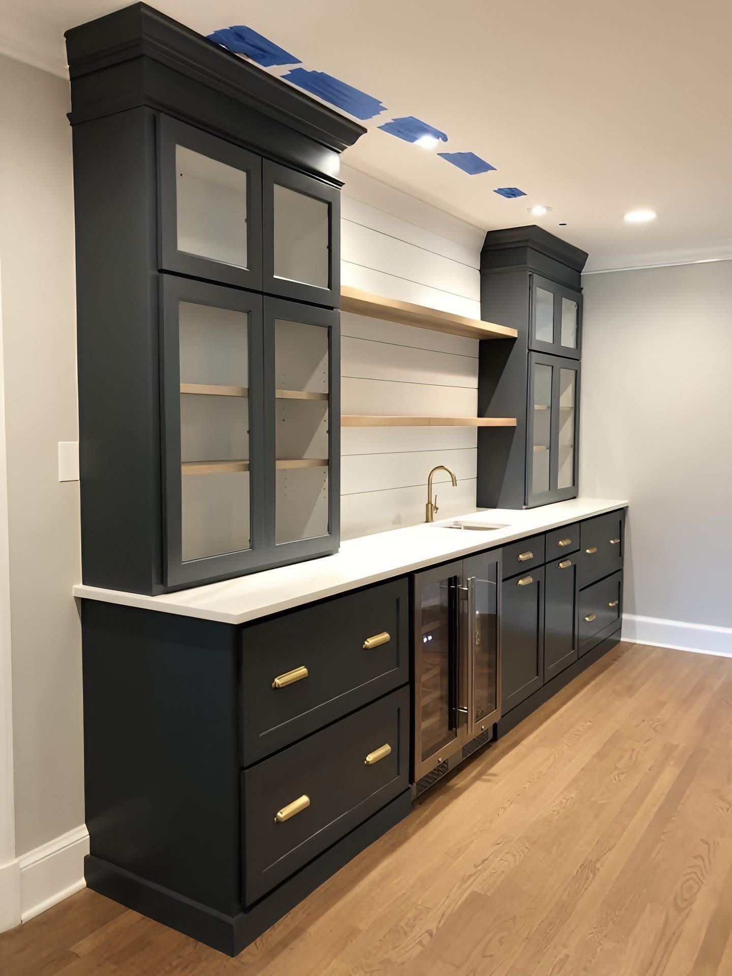 A home bar area with dark navy cabinets, glass-front upper storage, gold hardware, open wood shelving, and a wine fridge.