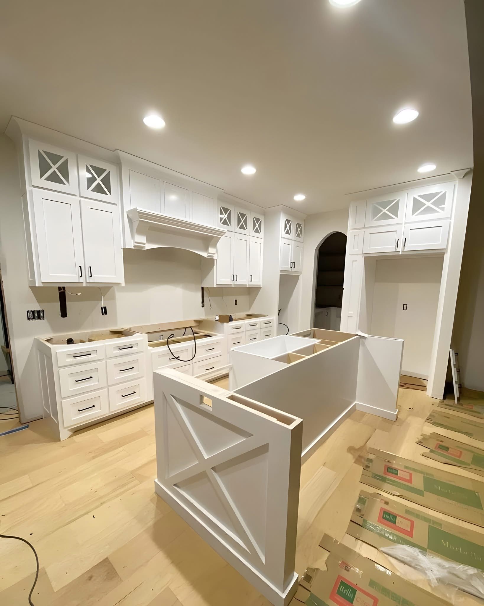A white kitchen under construction with an island, upper cabinets, and light wood flooring in a bright, modern space.