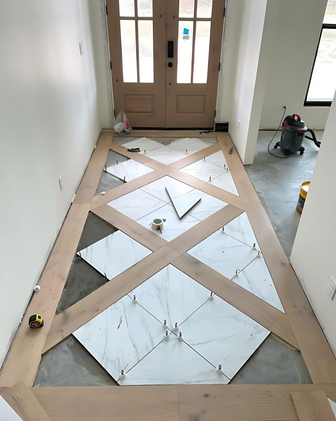 A foyer floor under construction featuring a geometric pattern of white marble tiles framed by light wood planks.
