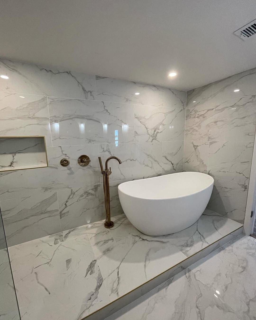 A modern white soaking tub sits on a raised platform tiled in white marble with grey veins, alongside a brass faucet.
