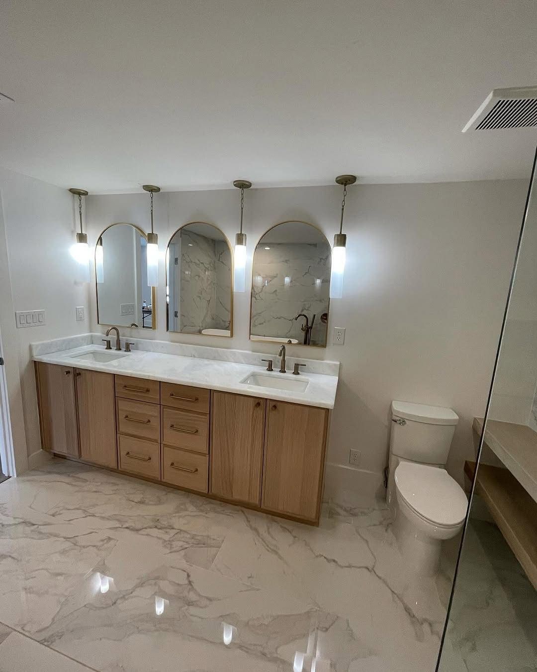 A modern bathroom with a light-wood double vanity, white marble countertop, three arched mirrors, and marble flooring.