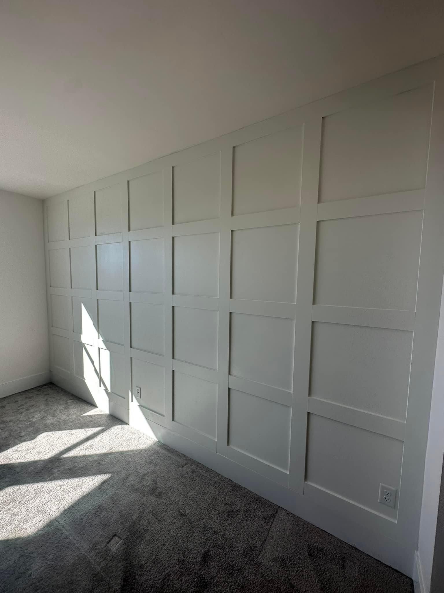 A wall featuring grid-style white wainscoting with square panels in a room with light gray speckled carpet.