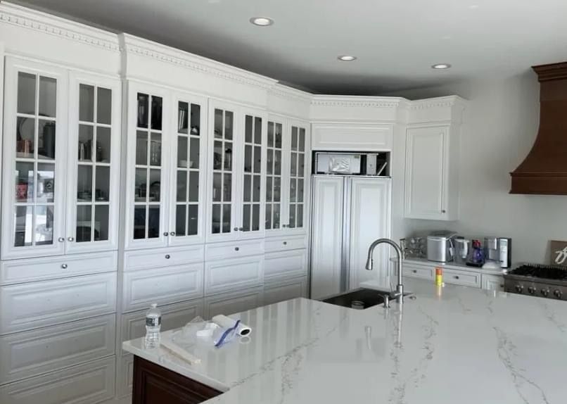 A modern white kitchen with floor-to-ceiling glass-front cabinetry, a large marble island, and a dark wooden range hood.