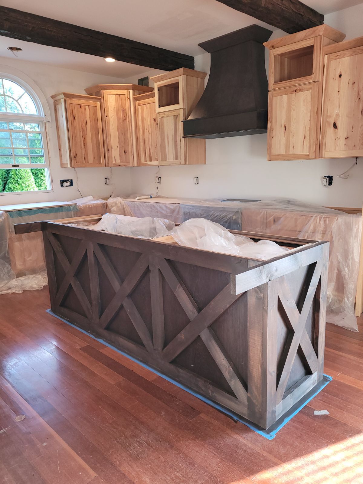 A kitchen under renovation featuring natural wood upper cabinets, a dark farmhouse-style island, and dark ceiling beams.