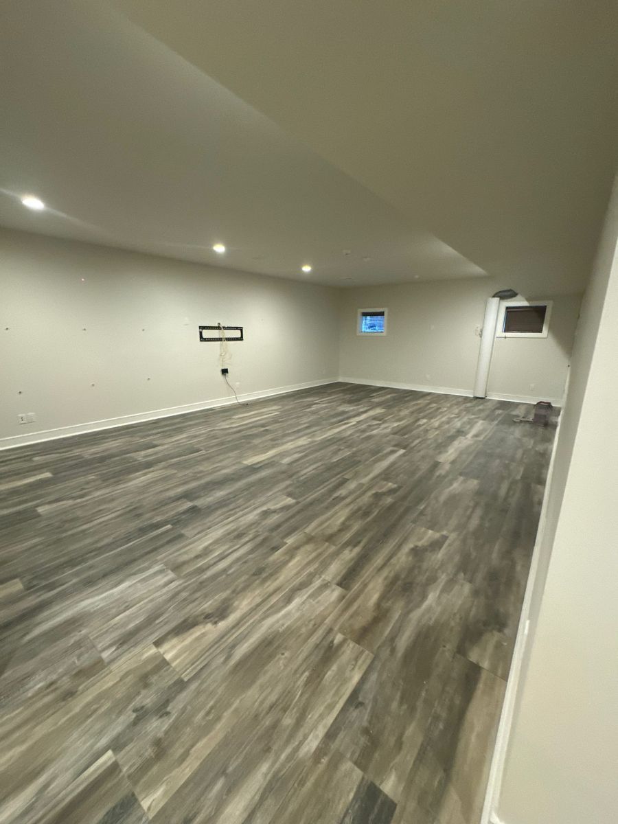A spacious, empty basement with light gray walls, white baseboards, recessed ceiling lights, and wood-look plank flooring.