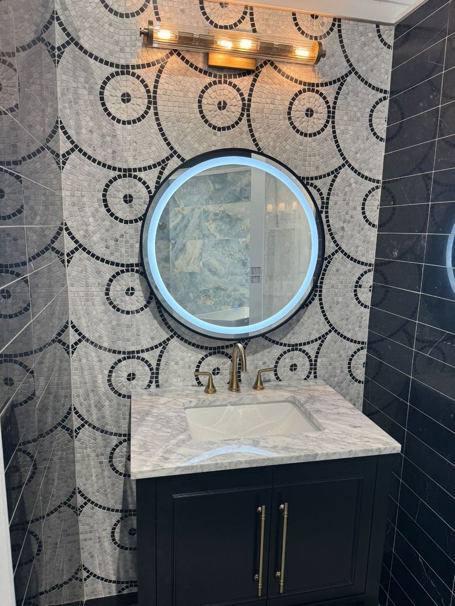 A bathroom vanity featuring a round LED-lit mirror, marble countertop, dark cabinet, and decorative circular wall tile.