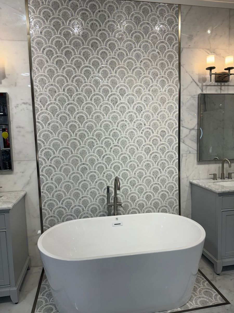 A freestanding white bathtub sits in front of a gray scalloped mosaic tiled feature wall in a modern bathroom.