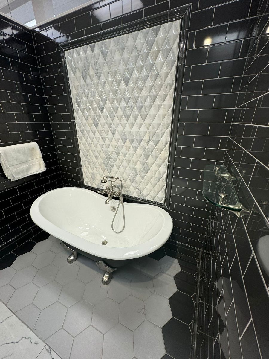 A white clawfoot bathtub sits against a black tiled wall with a central diamond-patterned white marble backsplash.