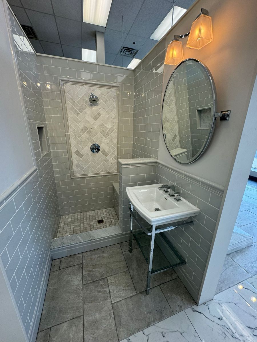 A modern bathroom vignette featuring a shower with herringbone tile, grey wall tiles, a pedestal sink, and a round mirror.