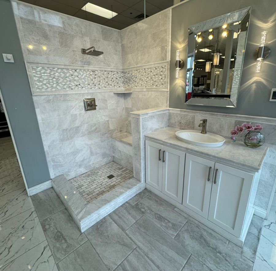 A modern bathroom display featuring a tiled walk-in shower with a bench and a white vanity with an oval sink and mirror.