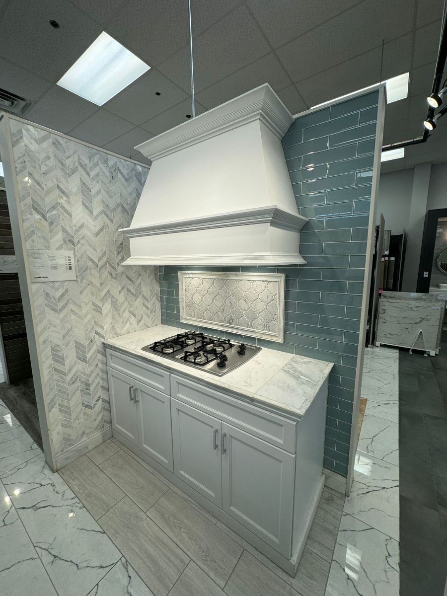 A kitchen display featuring white cabinets, a matching vent hood, a gas stovetop, and teal brick-style wall tiles.