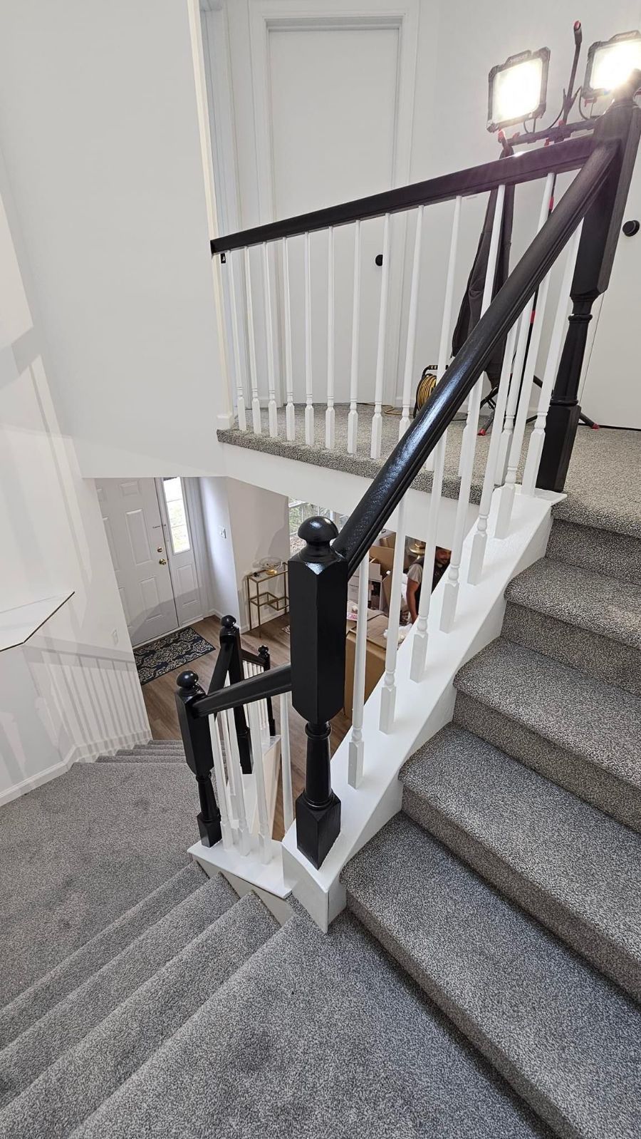 A staircase with gray carpet and a white railing with black posts, looking down from a landing into a brightly lit entryway.
