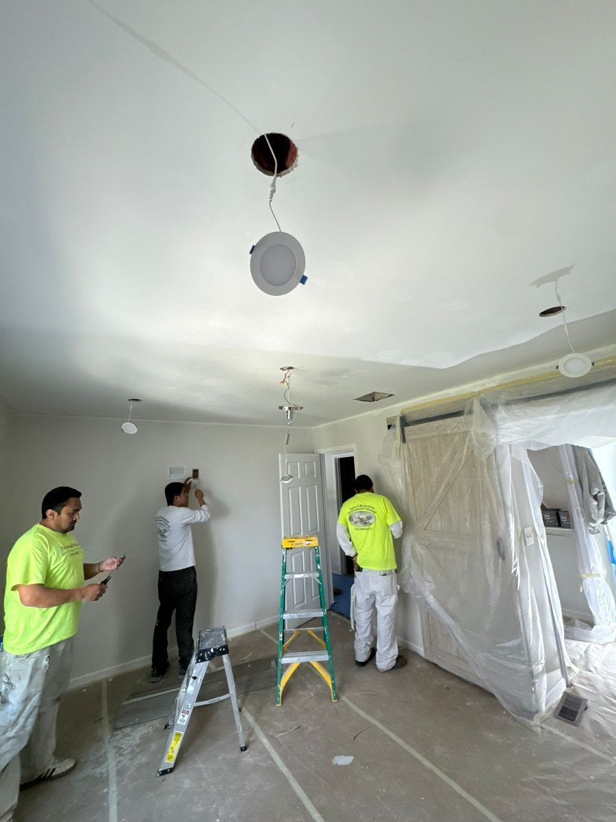 Three workers in neon yellow and white uniforms perform construction and electrical work inside a room draped in plastic.