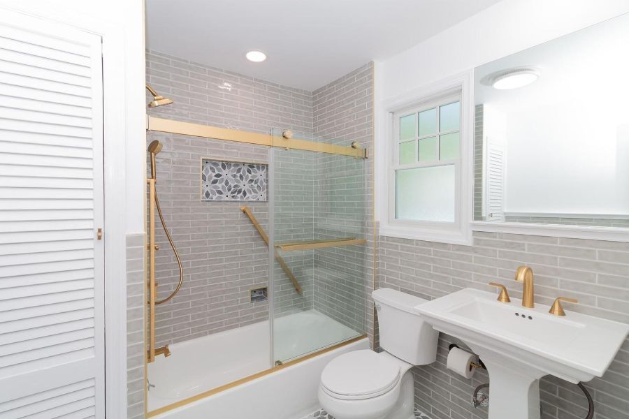 A bathroom featuring a shower with a sliding glass door, grey subway tile, a white pedestal sink, and a white toilet.