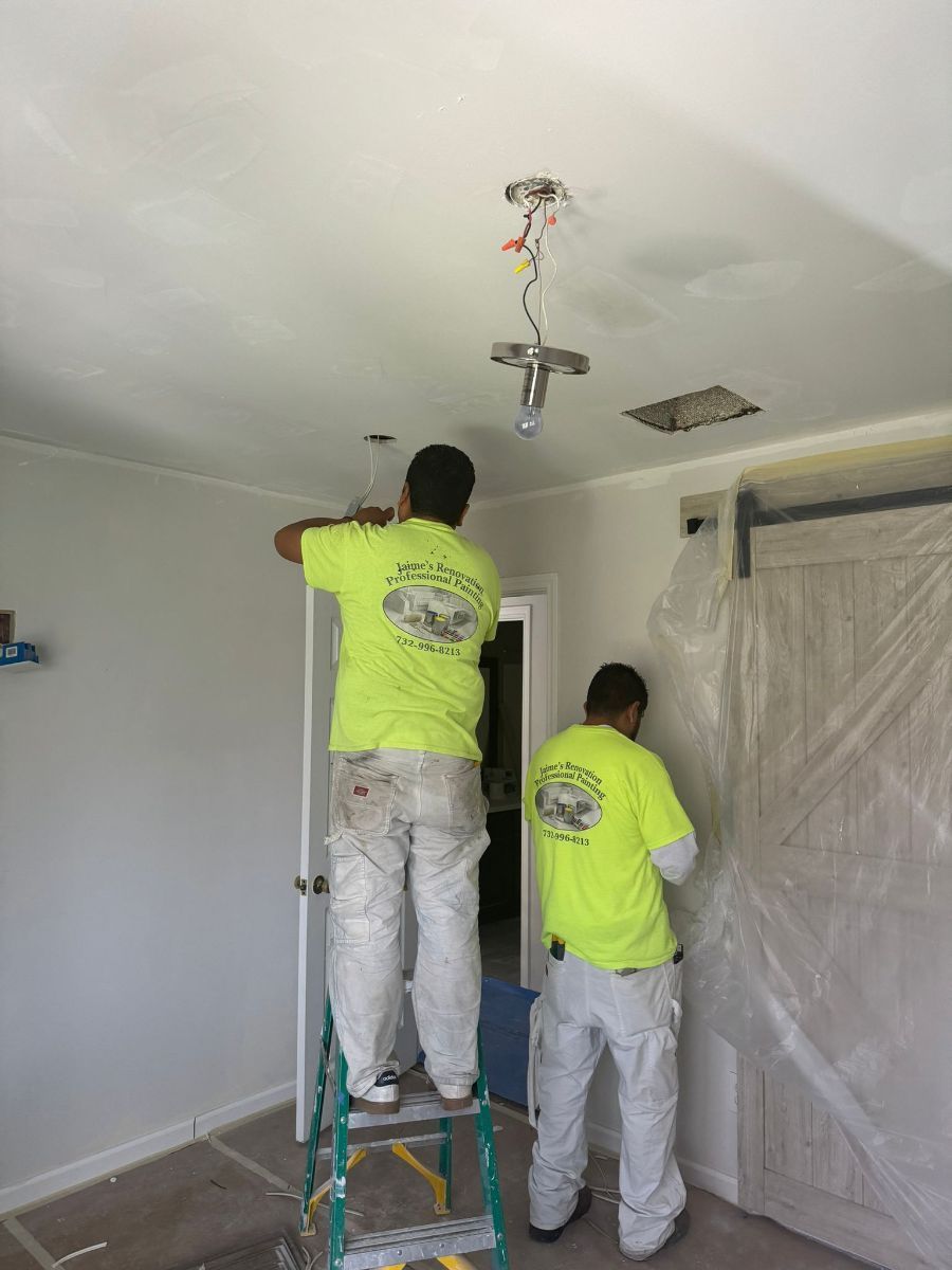 Two workers in high-visibility yellow shirts perform ceiling repairs inside a room covered in plastic protective sheeting.