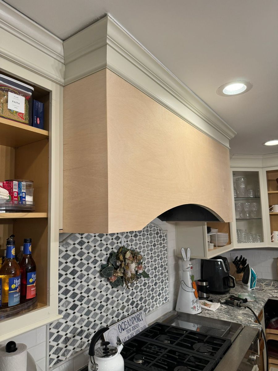 A kitchen range hood with light wood paneling, decorative crown molding, and a patterned tiled backsplash above the stove.