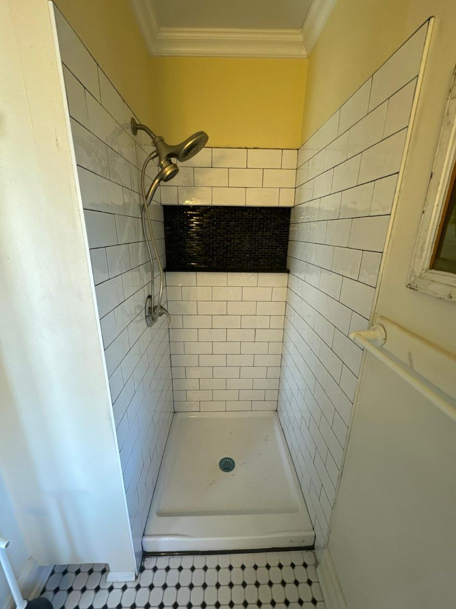 A walk-in shower with white subway tiles, a black tiled accent row, a brass shower head, and a checkered floor.