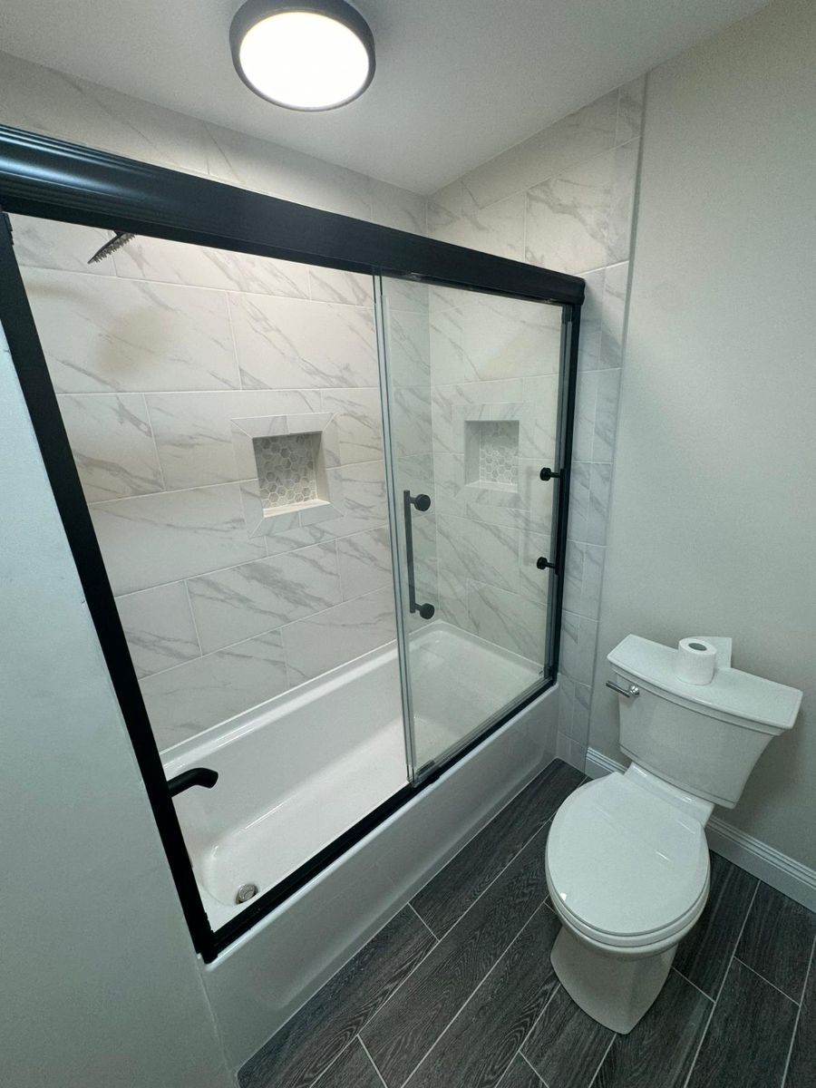 A modern bathroom featuring a white bathtub with a black-framed sliding glass door, marble-style tile, and a white toilet.