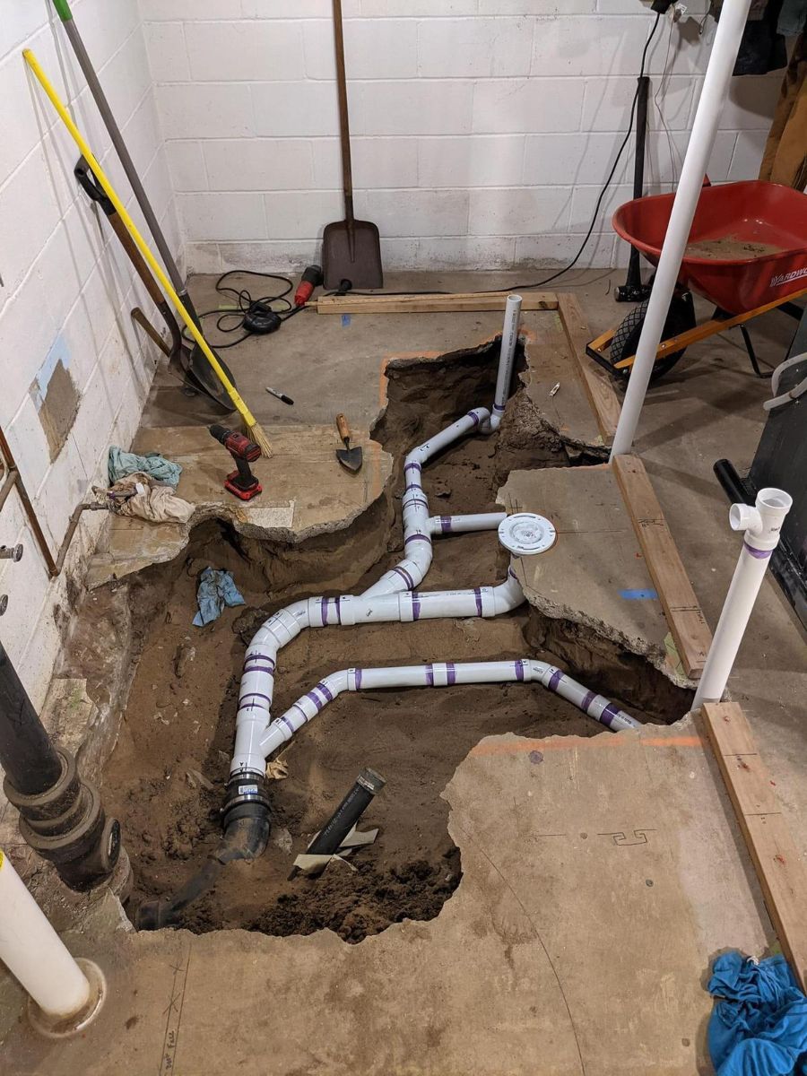 A trench dug into a concrete basement floor reveals newly installed white PVC plumbing pipes connected to a main drain.