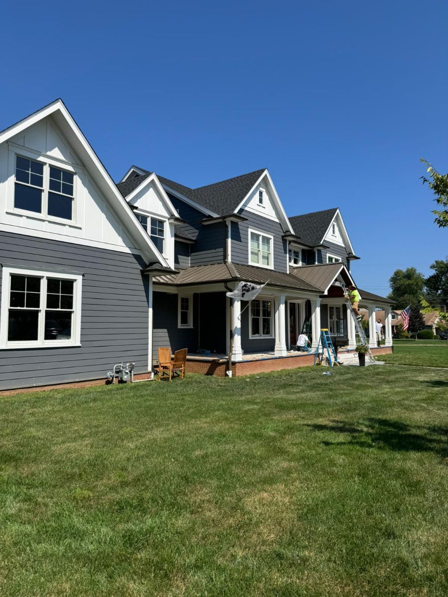 A large, grey-sided house with a porch and white trim stands under a clear blue sky, with workers present near the porch.