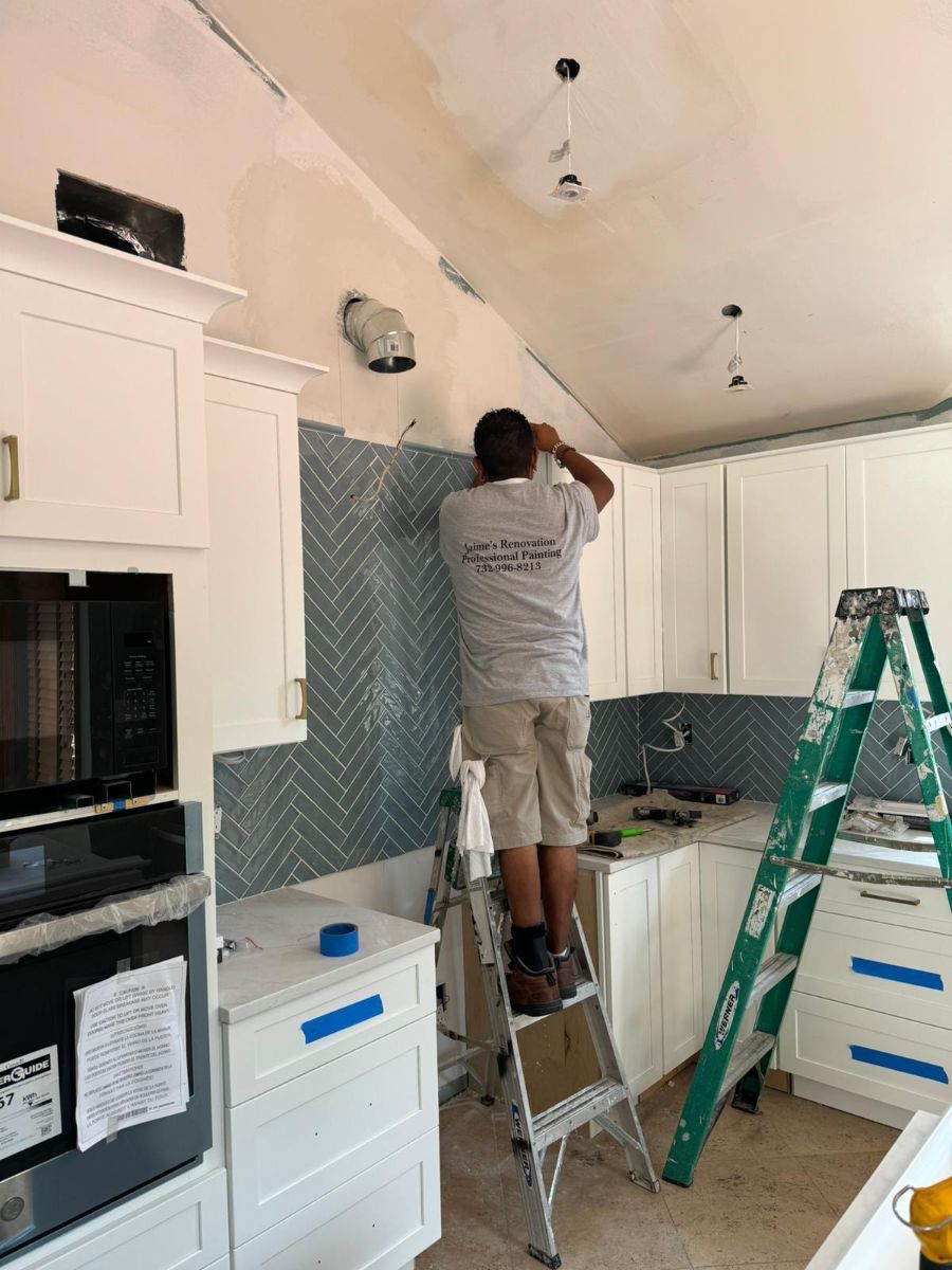 A worker on a step ladder installs gray, herringbone-patterned tile on a kitchen backsplash between white cabinets.
