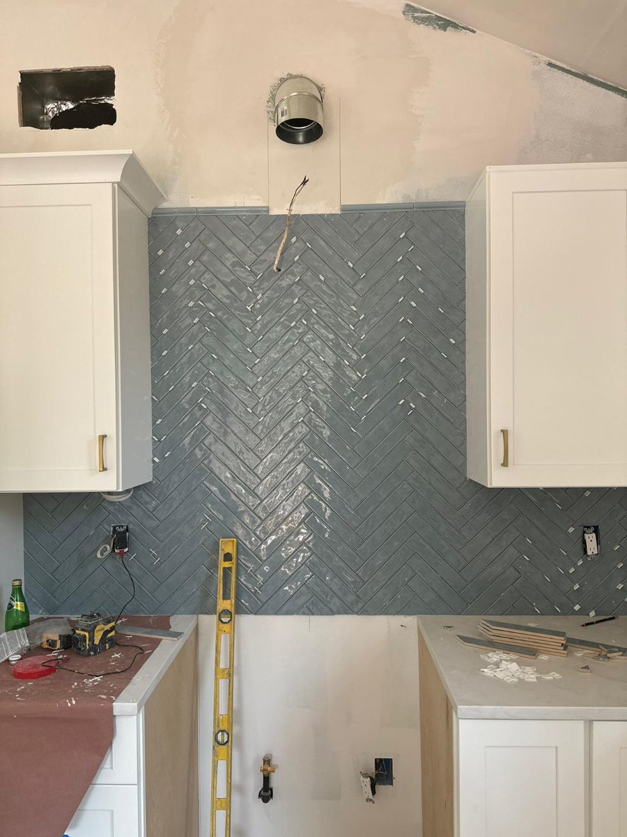 Kitchen under construction with white cabinets and a gray herringbone tile backsplash centered on a wall.