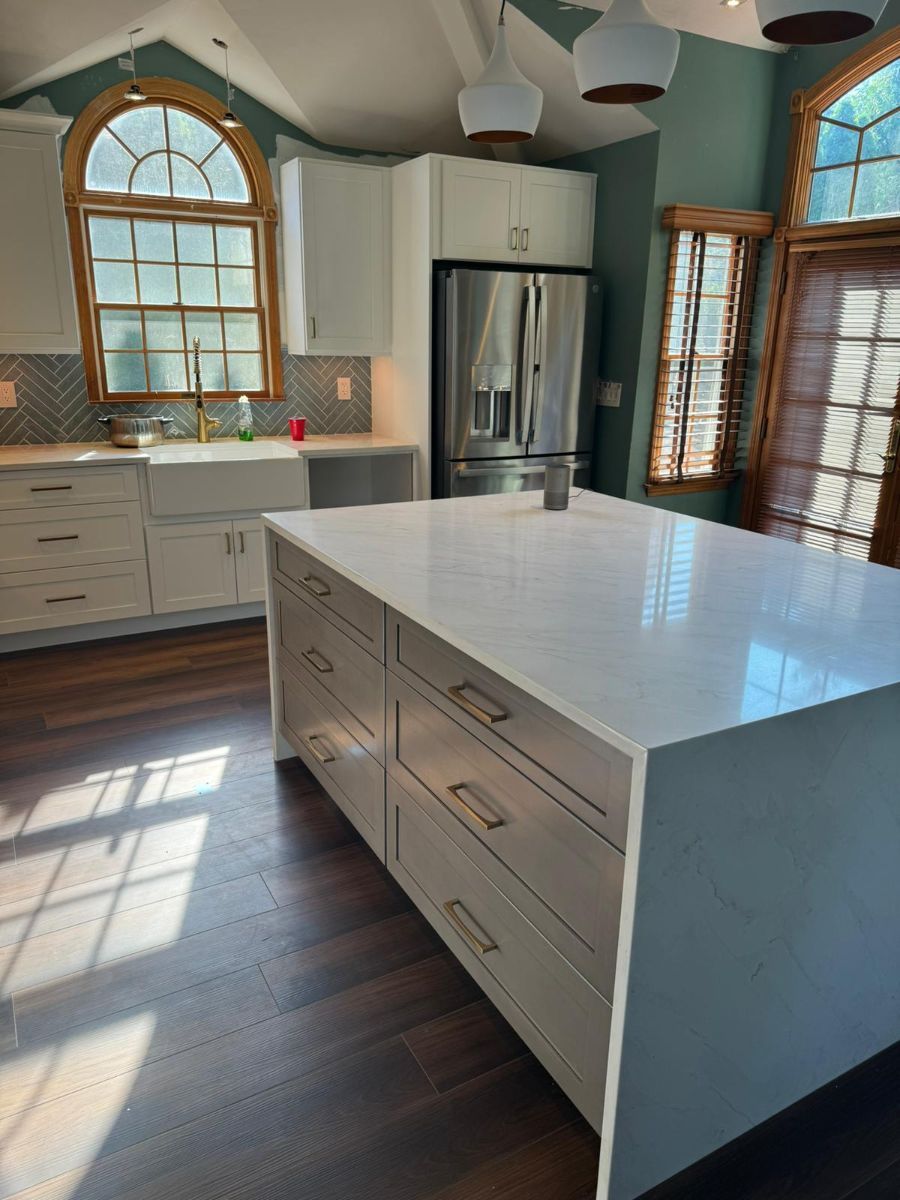 A modern kitchen with white cabinets, a large white kitchen island with drawers, and dark wood floors under arched windows.