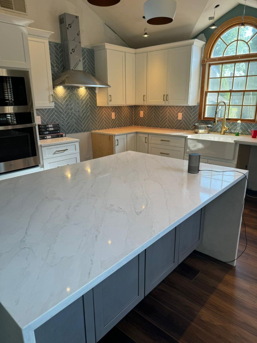 A modern kitchen featuring a large white quartz island, gray base cabinets, white upper cabinets, and herringbone backsplash.