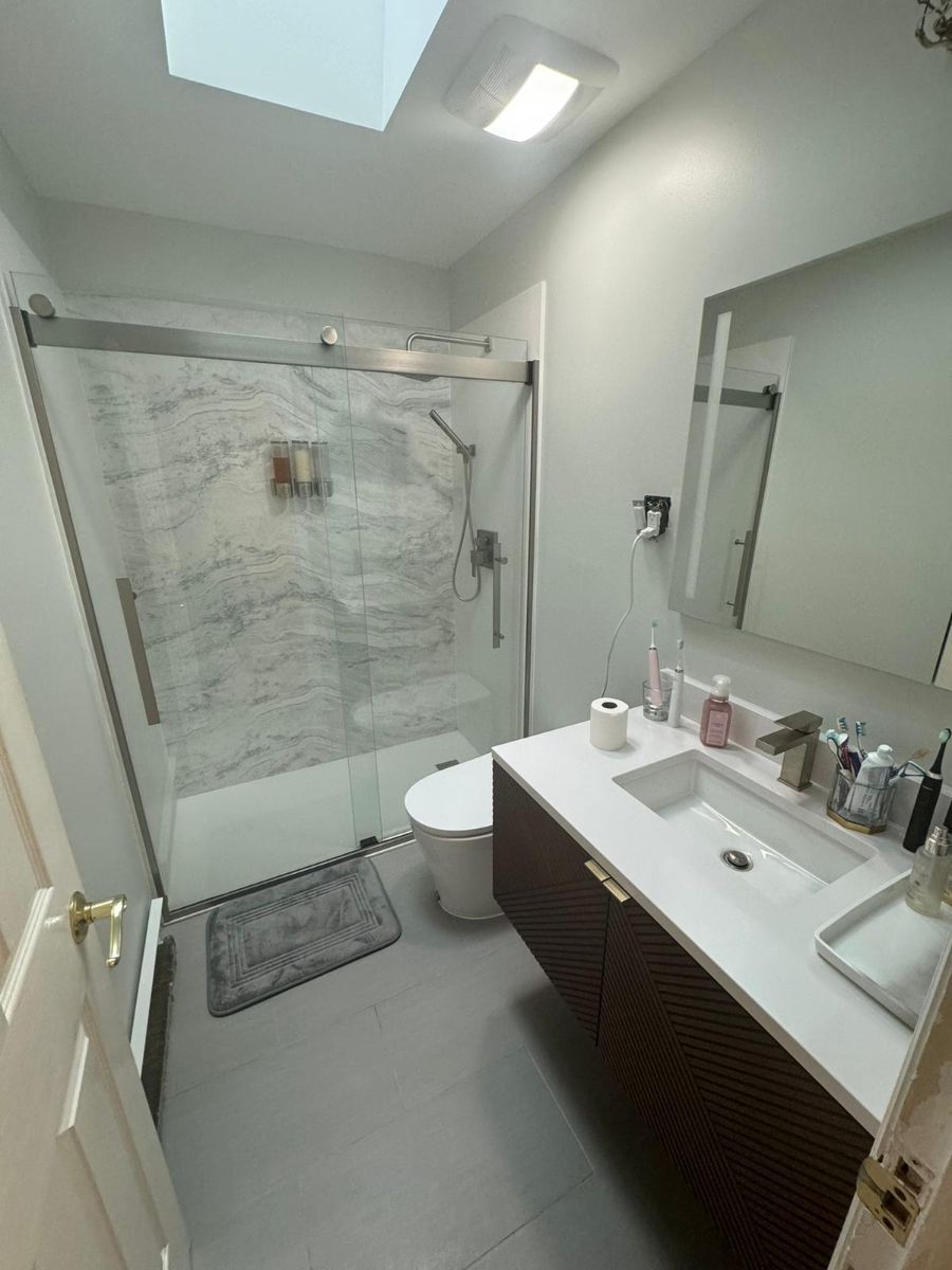 A modern bathroom with a glass-enclosed shower, marble tile walls, a dark wood floating vanity, and a skylight above.