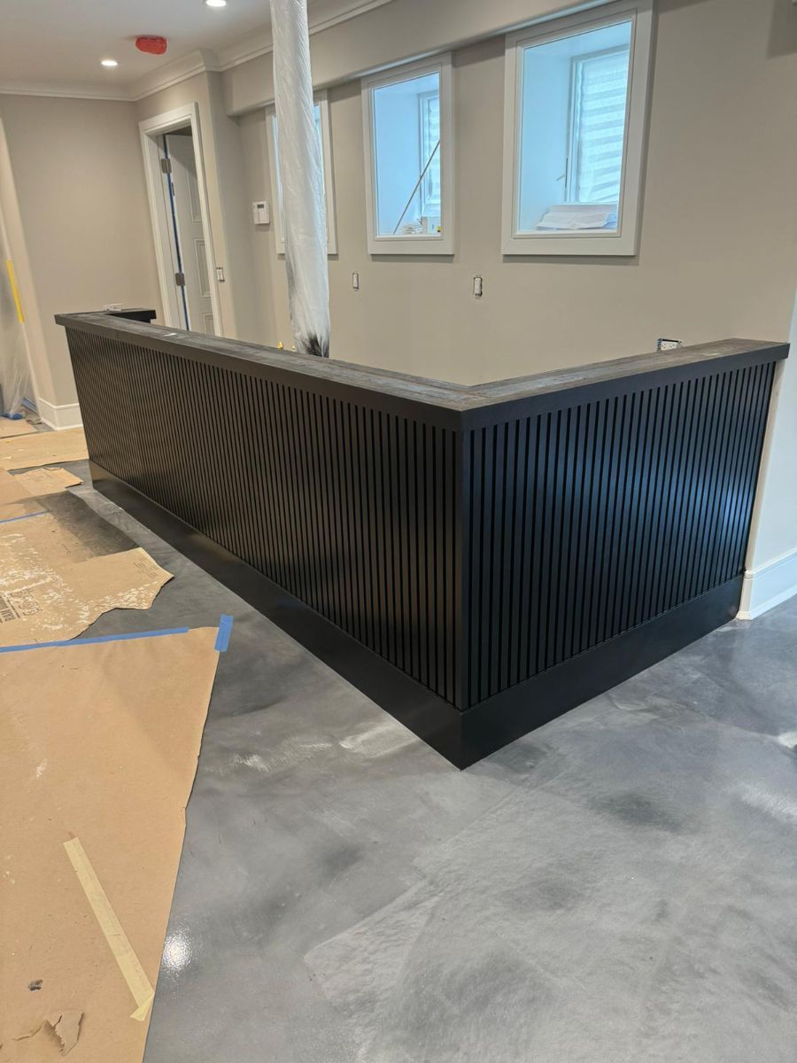 A black L-shaped reception desk with a textured, wavy slatted front stands in a room with light grey walls and concrete floors.