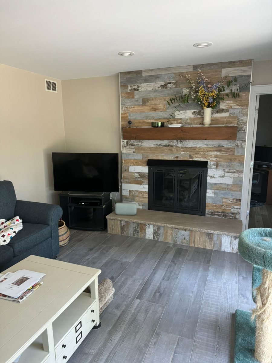 A living room featuring a stone fireplace with a wood mantel, a television on a stand, and light wood-look flooring.