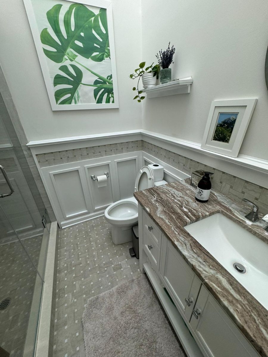 A modern bathroom with a white vanity, marble-patterned countertop, toilet, wood-paneled walls, and botanical wall art.