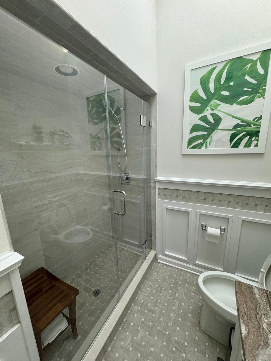 A modern bathroom featuring a glass-enclosed shower with a teak bench, gray tiled flooring, and a toilet with wainscoting.