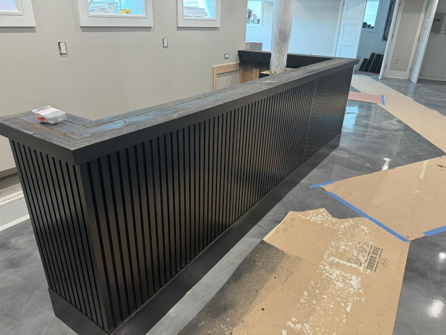 A dark wood reception desk with a vertical slat front stands in an unfinished room with a polished concrete floor.