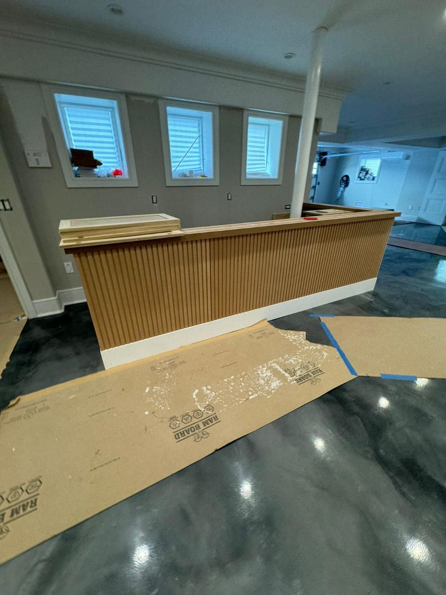 A light wood-slatted bar counter sits in a basement with gray walls, white trim, and a polished gray epoxy floor.
