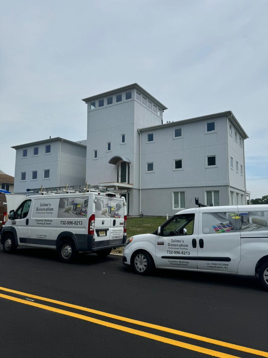 Two white work vans parked in front of a three-story, light-gray building with a central tower under a cloudy sky.