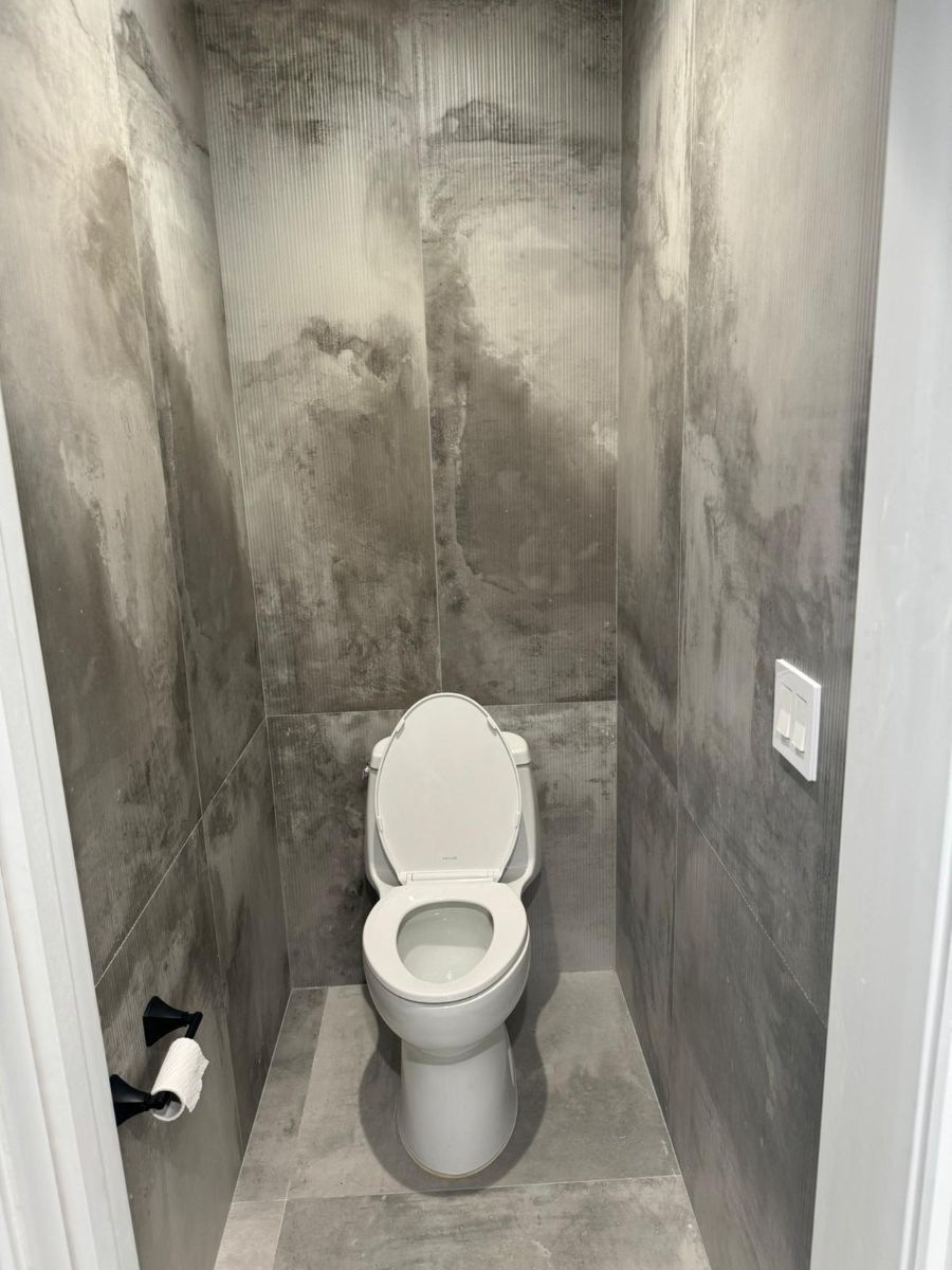 A small, narrow toilet room with gray, mottled, large-format tile walls and a white toilet with a black toilet paper holder.