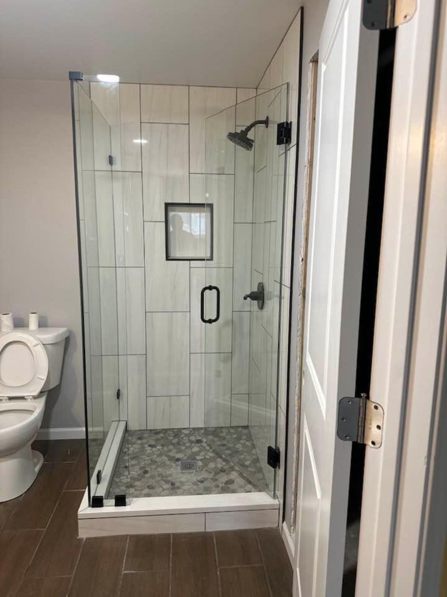 A corner glass walk-in shower with vertical white tile walls, a dark stone floor, and a small recessed niche in a bathroom.