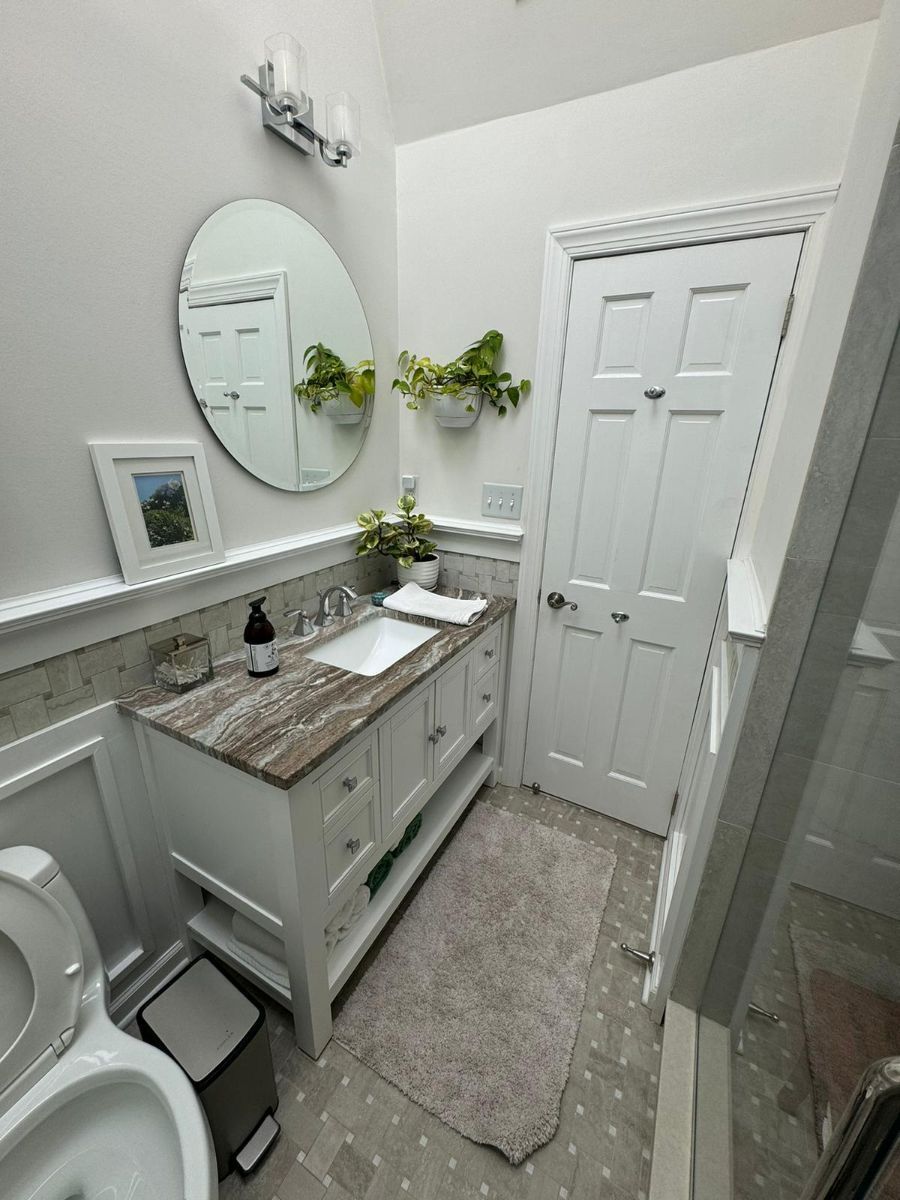 A bright bathroom with a white vanity, marble countertop, round mirror, potted plants, and a closed white door.