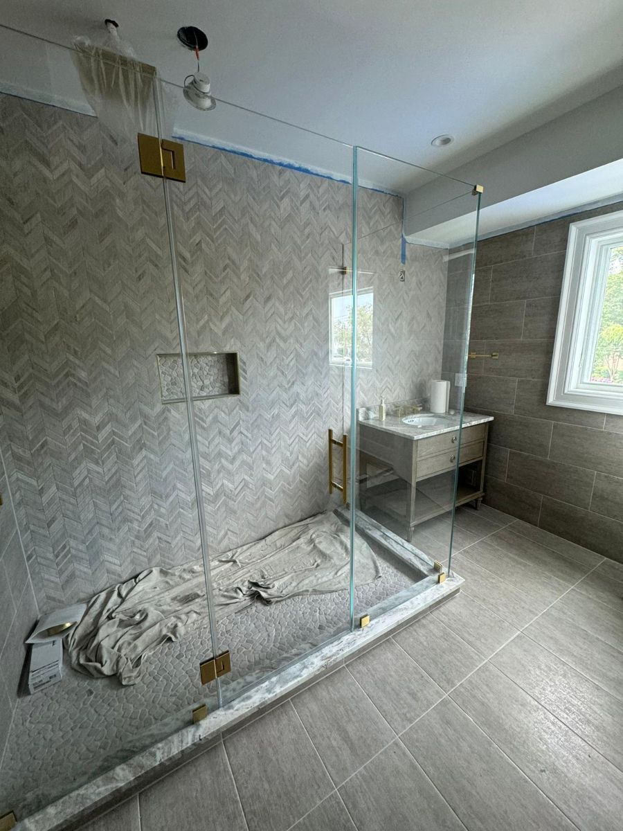 A modern walk-in shower with light grey herringbone wall tiles, a glass enclosure, and a cloth lying on the shower floor.