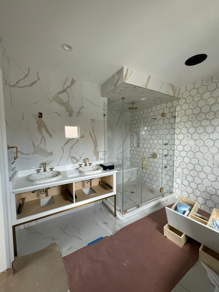 A modern bathroom under construction with dual sinks on a white marble vanity, a glass shower, and hexagonal wall tiles.